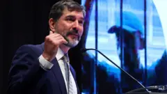 Daniel Johnson vestido de terno e gravata, falando a um microfone e gesticulando com a mão direita