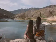 Delal (à esquerda) faz uma pausa com uma companheira de combate perto de um pequeno lago próximo à sua base. Elas estão de costas para a câmera e vestem trajes tradicionais curdos.