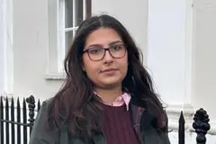 Maryam Khan está de pie afuera, junto a un edificio con una verja de hierro forjado. Sonríe a la cámara y lleva gafas, una camisa rosa con cuello, un suéter bordó y una chaqueta verde.