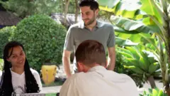 Três pessoas, dois homens brancos e uma menina morena, estão reunidas em torno de uma mesa, com plantas e arbustos verdes ao fundo
