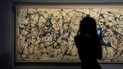 A person with long hair in silhouette is seen facing the artwork holding their mobile phone to video the painting. The artwork is long horizontally and the grey canvass appears a light tan colour under the gallery lights. It features black paint flicks and splashes all over it, together with some fewer red markings.