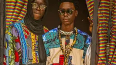 Two men in multi-patterned colourful patchwork clothes dey look camera by a doorway. One wear a black headscarf and glasses, di oda dey in sunglasses and get beaded necklaces around im neck - for Diourbel, Senegal, April 2024