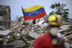 En la fotografía, tomada en Caraballeda, Venezuela, el 20 de julio, se ve una bandera venezolana sobre un montón de escombros, con una palmera al fondo y un cielo azul de fondo, mientras un rescatista camina en primer plano con efecto bokeh.