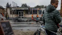 Мальчик в зелёной куртке с велосипедом на фоне разрушенного здания и сгоревшего автомобиля. В кадре видны спасатели в жёлтых и оранжевых жилетах