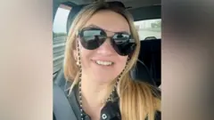 Polina Azarnykh olha de frente para a câmera, sentada em um veículo. Ela tem longos cabelos loiros, usa óculos de sol e está sorrindo.