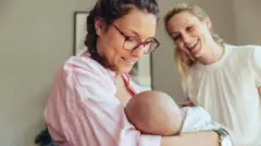 Una mujer con su bebé en brazos y otra que la mira sonriente.