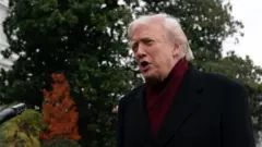 暗い色のコートにえんじ色のマフラーをしたトランプ氏が、記者団のマイクを前に話している。後ろの木々の一部が紅葉している