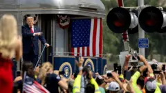 Трамп стоит в хвосте поезда с американским флагом, перед ним — много людей.