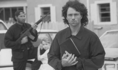 David Koresh tient un livre, un homme armé d'un pistolet automatique et un petit enfant sont à l'arrière-plan.