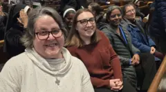 Freiras estão sentadas lado a lado, vestindo roupas comuns e sorrindo para uma câmera posicionada ao lado. A religiosa mais próxima da câmera usa um pequeno crucifixo prateado no pescoço. Ao fundo, outras freiras aparecem sentadas com hábitos tradicionais, mas seus rostos não são visíveis
