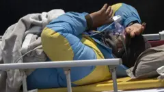 A man in a blue and yellow jacket shields his eyes with his arm as he lies on a yellow emergency stretcher under a grey blanket.