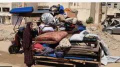 Fọto yii ṣafihan iya agba kan to duro ti awọn ẹru rẹ nigboro Gaza, iya naa n wo bii ẹni ti ọrọ su. Awọn ẹru bii ibusun, apo loriṣiriṣi ni iya naa to pọ, ti ko jọ pe o mọ ibi to fẹẹ ko wọn lọ. Aworan ile to ti wo wa lẹyin rẹ ati awọn biriki afọku gbogbo.