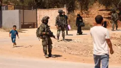 迷彩柄の軍服とヘルメット、顔を隠すマスクを着用し、ライフルを携えたイスラエルの兵士3人が、乾燥した丘の道を歩いている。兵士たちの周りには、男性2人、女性1人、少年1人の計4人のパレスチナ人住民が歩いている