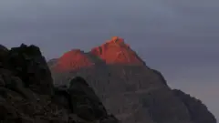 O pico do Monte Sinai ao anoitecer, em 2024. A luz ilumina o topo da montanha rochosa, que se eleva acima de outra montanha em primeiro plano.