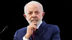 Lula veste terno azul escuro e gravata roxa, com a mão no queixo. Ele parece observar alguém enquanto fala num palco.