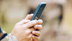 Ibiganza bibiri bifashe telefone igendanwa, hagaragara n’umwambaro wa gisirikare ku kuboko. 