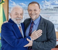 Foto de Edinho Silva e Lula apertando as mãos e se abraçando em um gabinete com uma bandeira do Brasil ao fundo