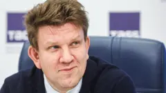 Евгений Капьев в костюме с галстуком на фоне спинки кожаного кресла и логотипов ТАСС.