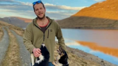 Homem com óculos de sol apoiados na cabeça sorri para a câmera, enquanto dois cães preto e branco pulam ao seu redor