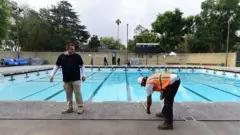 Funcionário faz medição na borda de uma piscina, observado por outra pessoa