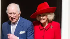 Król Karol uśmiecha się i patrzy prosto przed siebie. Ma na sobie granatowy garnitur w prążki, białą koszulę, srebrny krawat oraz srebrną poszetkę. Jego lewa dłoń jest lekko uniesiona, a na nadgarstku ma czarno‑złoty zegarek. Na małym palcu lewej dłoni nosi duży złoty pierścień.
Po jego lewej stronie stoi królowa Kamila. Ma na sobie jaskrawoczerwoną marynarkę z broszką w kształcie krzyża oraz czerwony kapelusz dopasowany kolorystycznie do stroju. Ona również patrzy prosto przed siebie i uśmiecha się