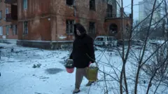 Жінка несе відра з уламками на тлі пошкодженого обстрілом будинку, на землі лежить сніг