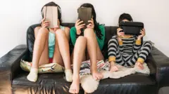 Três crianças sentadas em um sofá com o rosto grudado em telas