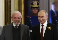 Lula sonriente camina junto a Vladimir Putin. Lula lleva una chaqueta de traje gris oscuro, con un chaleco oscuro, una camisa blanca y corbata. Putin luca una chaqueta oscuro en cuya solapa tiene un prendedor con rayas amarillas, junto a una camisa blanca y una corbata vino tinto.