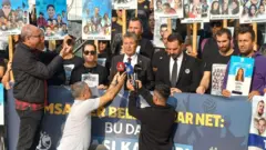 Kalabalık bir grup pankartalarla sesiz duruyor, bir erkek mikrofonlara konuşuyor