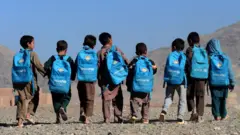 چند کودک که بیگ‌های مکتب با نشانی یونیسف در پشت شان است در حال راه رفتن هستند.