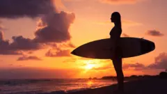 Mujer con una tabla de surf en un atardecer.