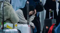 Seorang perempuan muda berhijab sedang duduk dengan wajah lesu dan mata terpejam dengan tangan kirinya memegang kepala di tengah banyak orang.