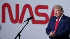 O presidente dos EUA, Donald Trump, veste terno azul-escuro e gravata vermelha enquanto fala ao microfone em um palco com um grande letreiro da Nasa ao fundo, em letras vermelhas, em 30 de maio de 2020