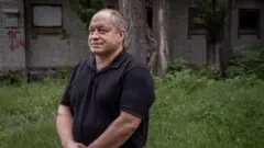 El doctor Serhii Andriichenko se encuentra en una zona cubierta de césped, vistiendo un polo negro y con las manos entrelazadas frente a él.