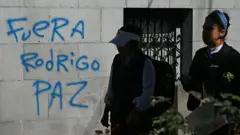 Mulheres caminham ao lado de um grafite que diz "Fora Rodrigo Paz", em referência ao presidente da Bolívia, em La Paz, em 19 de maio de 2026.