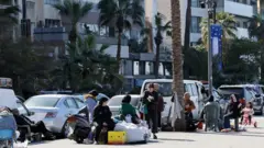 Saldırıdan kaçan insanlar kaldırımda oturuyor yanlarında araçlar park etmiş.