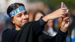 Mulher jovem com uma faixa na testa escrita Jesus tirando selfie com o celular durante a Marcha para Jesus em maio de 2024