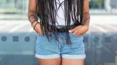 Дівчина тримає руки в кишенях коротких джинсових шортів, її обличчя не в кадрі, вона має довгі тоненькі кіски