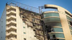 مبنى شاهق وحديث بيج اللون، تعرضت واجهته للتلف، الزجاج في بعض النوافذ في الطوابق العليا مهشم.
