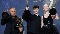 Stephen Graham, Owen Cooper y Erin Doherty, actores de la serie Adolescencia, muestran sus premios Emmy. Graham y Cooper visten trajes negros, mientras que Doherty, a la izquierda de la imagen lleva un vestido negro.