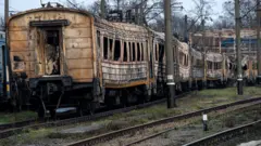Довгий ряд сильно пошкоджених і обгорілих вагонів електрички, що стоять на залізничних коліях. Корпуси вагонів почорнілі, з вибитими або деформованими вікнами, видно сліди пожежі та руйнувань по всій довжині складу.