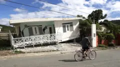 Una persona en bicicleta pasando frente a una casa dañada por el terremoto.