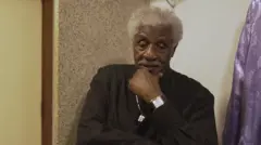 Tony Tornado é um homem negro, de cabelos brancos. Ele está com a mão no queixo