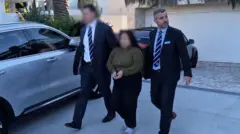 Two men in dark suits holding a handcuffed woman by the elbows in front of a house with two parked cars in the background