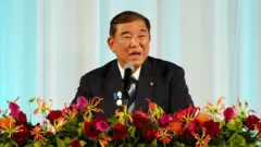 Премьер-министр Японии Сигэру Исиба сидит за столом перед микрофоном. Перед ним виден ряд ярких красных и розовых цветов