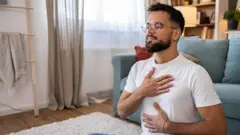 Homem fazendo exercícios de respiração, sentado no chão de uma sala, com uma mão no peito e a outra no abdômen