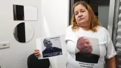 Antônia Regina segura foto do filho e usa camisetabetsport comhomenagem a Denner com a frase "Justiça pelo meu filho Denner"