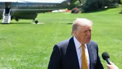 ABD Başkanı Donald Trump çimde mikrofona konuşuyor. Arkasında helikopter var.