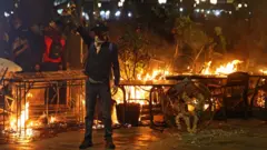 Демонстрант стоит с поднятым кулаком на фоне горящей баррикады