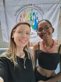 Fotografia colorida mostra duas mulheres de óculos, roupas pretas e sorrindo. Ao fundo, lê-se em um cartaz: "Diversidade Católica"